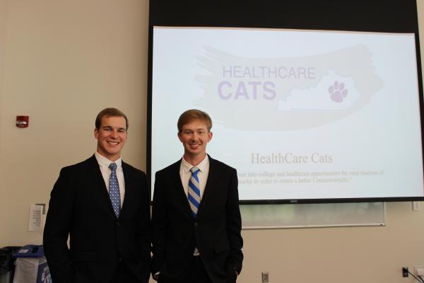 Photo of two members of healthcare cats in suits and ties, in front of a sign that says "Healthcare Cats."