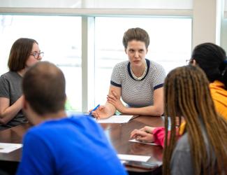 Woman speaking to a group of students at a conference table. Everyone at the table has a piece of paper and a pen.