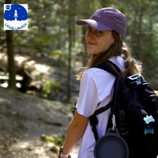 Megan Sutherland wearing a white t-shirt, a black backpack, and a purple baseball cap, hiking in the woods.