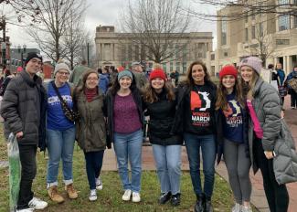 A group photo of 8 people who are part of the Lewis Student Diversity, Equity & Inclusion (DEI) Coalition at a women's march. It is a brisk, cold day as everyone in the photo is wearing thick coats and beanies.