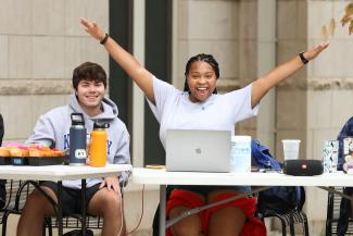 Two students sitting at some tables outside. The camera is focused on one student in a white shirt who has her arms out and smiling cheerfully at the camera. She has a laptop. The student on the left is smiling also.