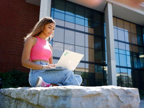 A girl is sitting cross-legged on a big rock outside of the Jacobs Science Building, looking at her laptop which is in her lap.