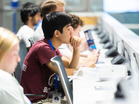 A group of students sitting in a lecture hall listening intently. The camera is focused on a boy in a red shirt and a blue lanyard.